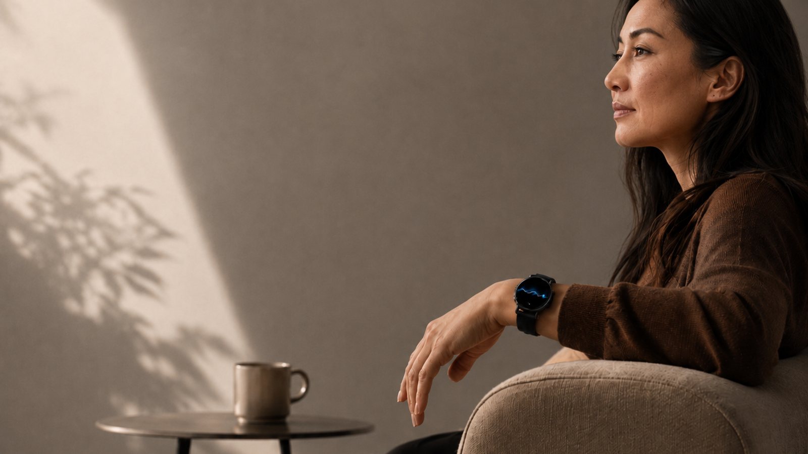 A woman relaxing in an armchair, her smartwatch quietly showing a pulse wave in warm daylight